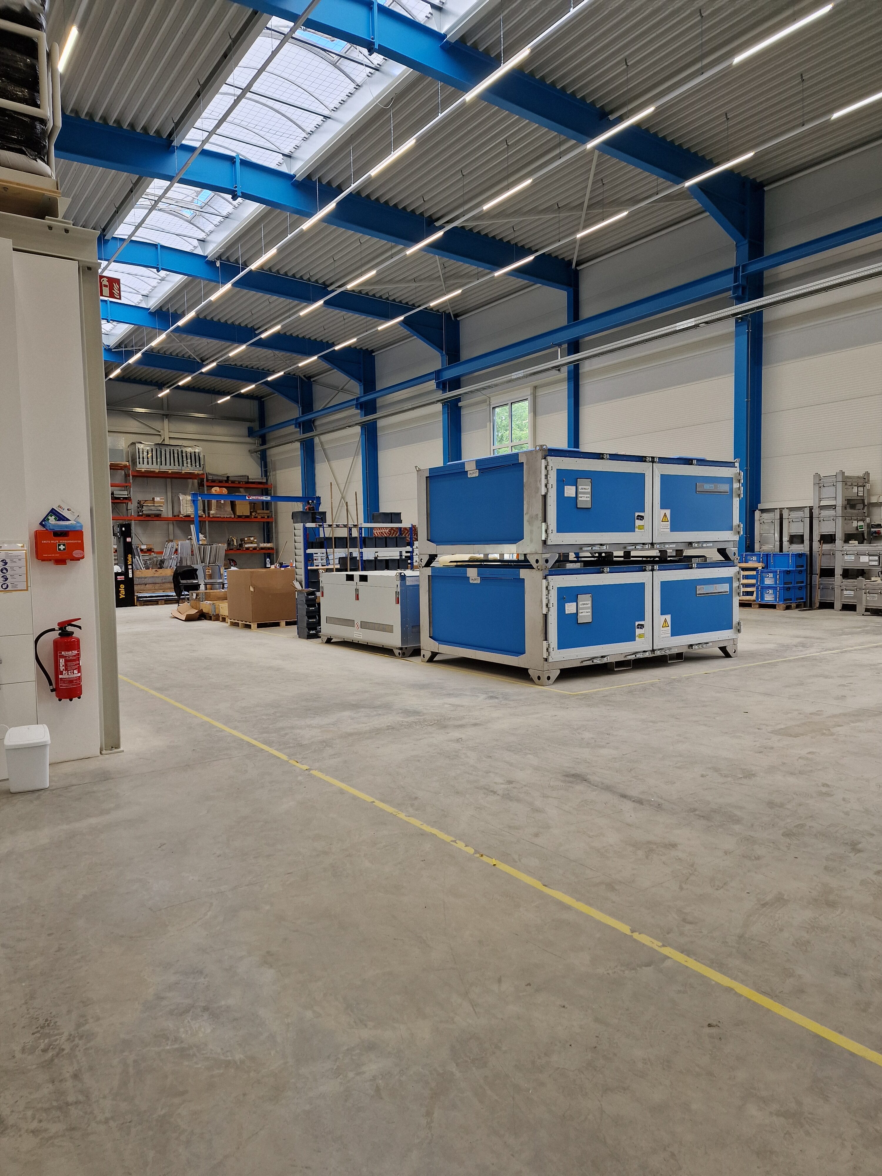 Große Lagerhalle mit blauen Containern, Regalen und Kartons, leere Bodenfläche. Helle Beleuchtung.